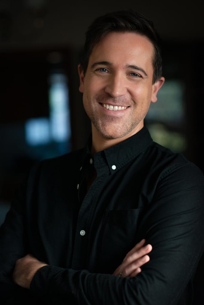 Photo of acting, accent and voice coach, Jack Starkey-Gill. He is smiling while crossing his arms and wearing a black, button up shirt with white buttons.